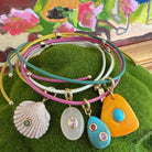A stack of five multi-strand leather wrap bracelets with gold-tone hardware and colorful pendants, displayed on a green textured surface with a painted floral background. The pendants include a white seashell, a milky white oval with a pink stone, a turquoise teardrop with a pink stone, a bright turquoise oval, and a large orange teardrop with a turquoise center.