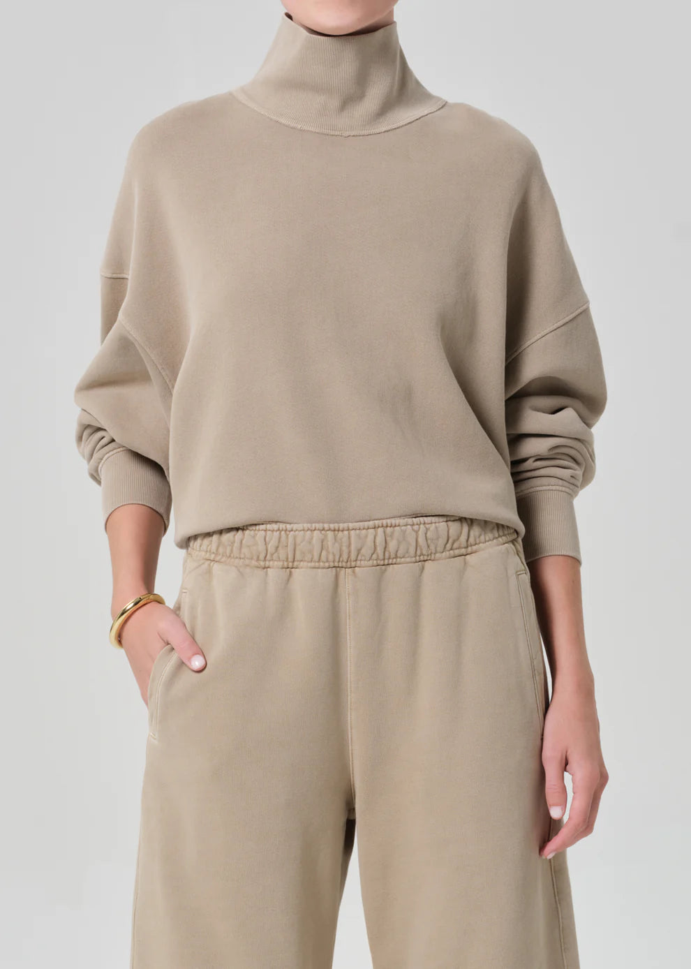 Person wearing a beige turtleneck sweater and pants on a light gray background