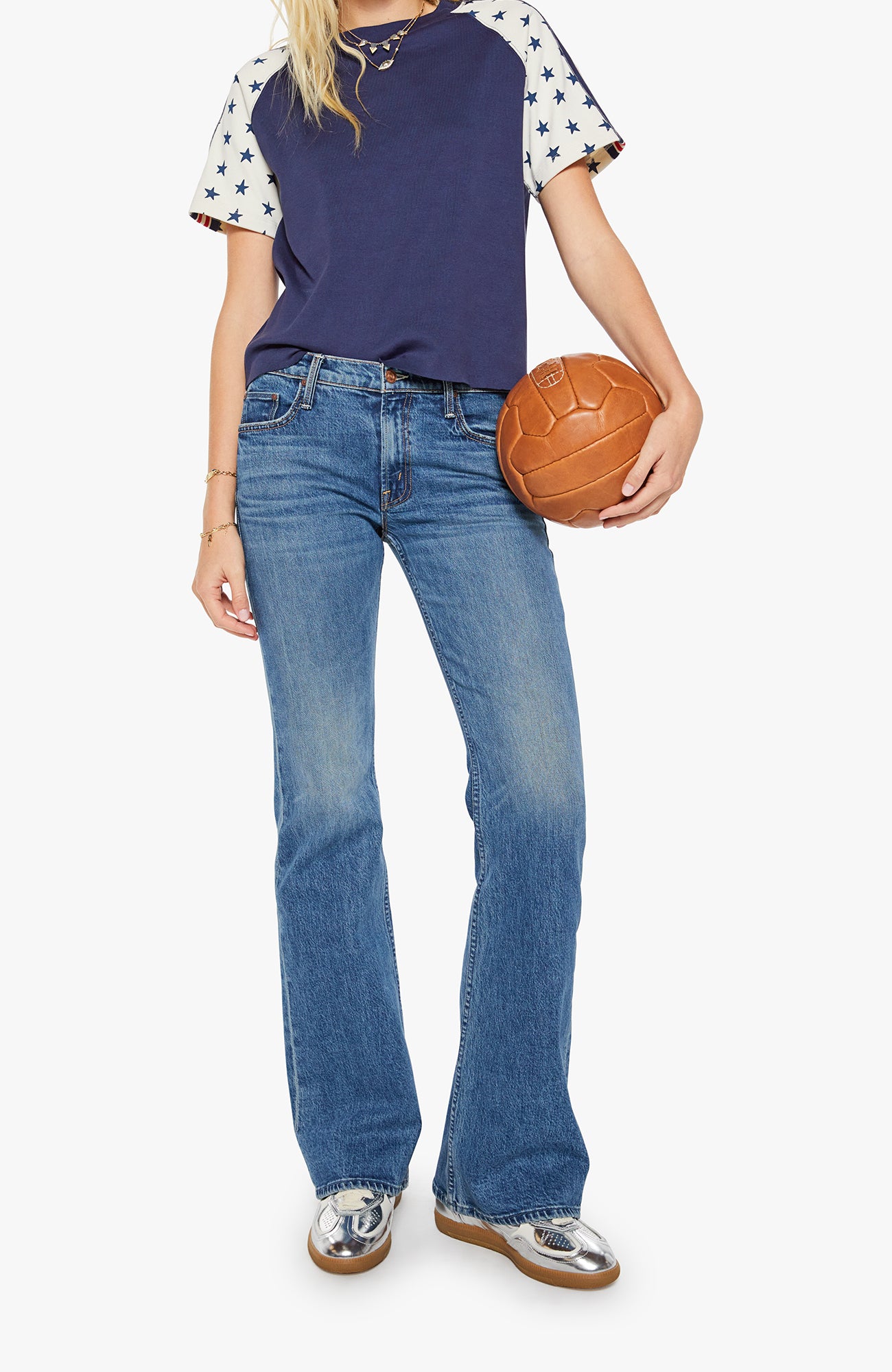 Person wearing a navy and white star-patterned shirt, blue jeans, and silver shoes, holding a brown leather ball.