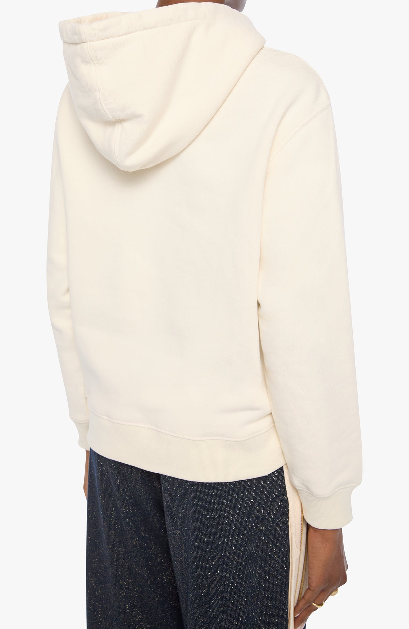 Back view of a person wearing a cream-colored hooded sweatshirt and dark charcoal pants against a plain white background.