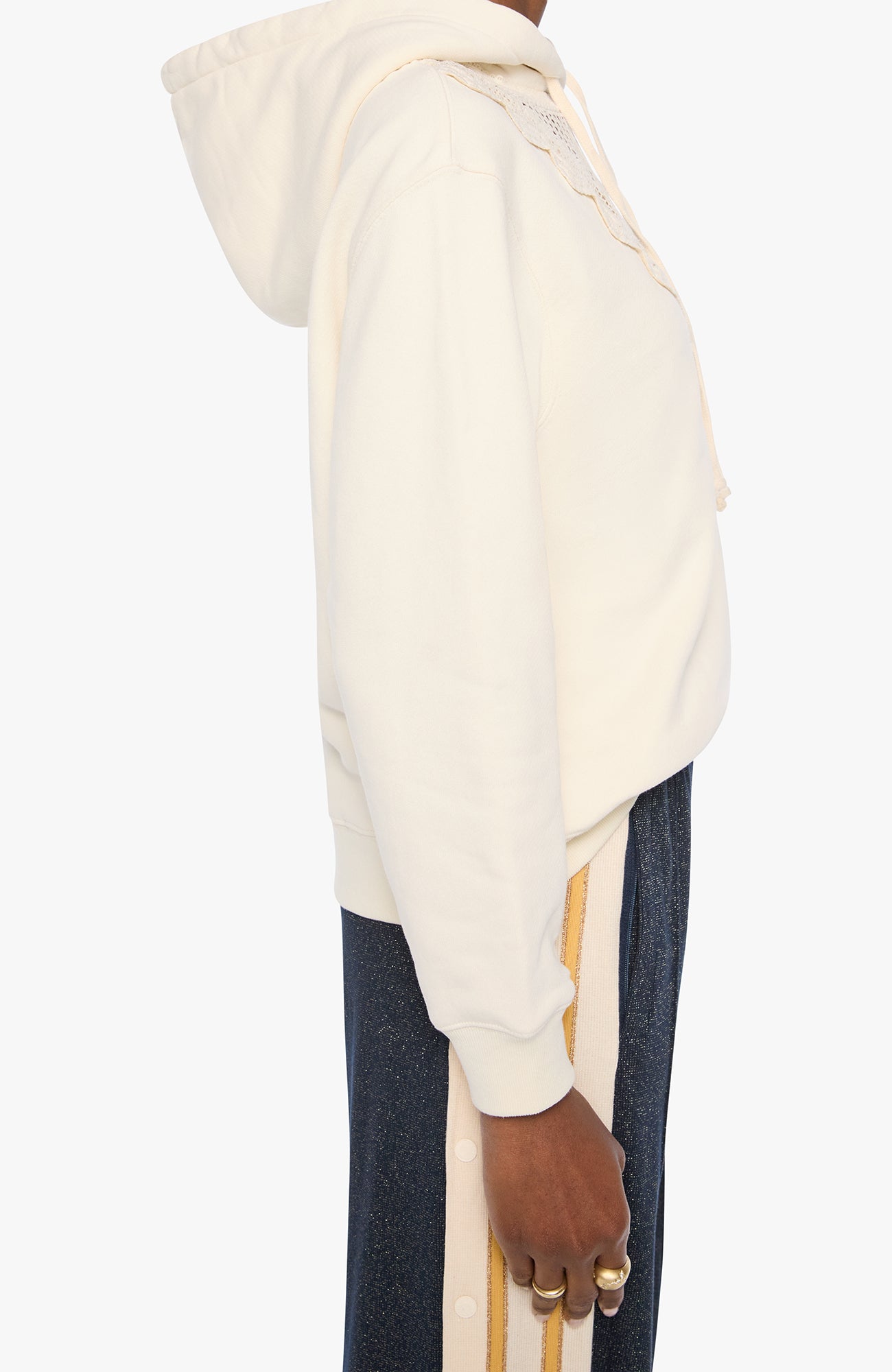 Side profile of a person wearing a cream-colored cropped hoodie with lace-up detailing on the hood and dark blue track pants with gold stripes, set against a plain light gray background.