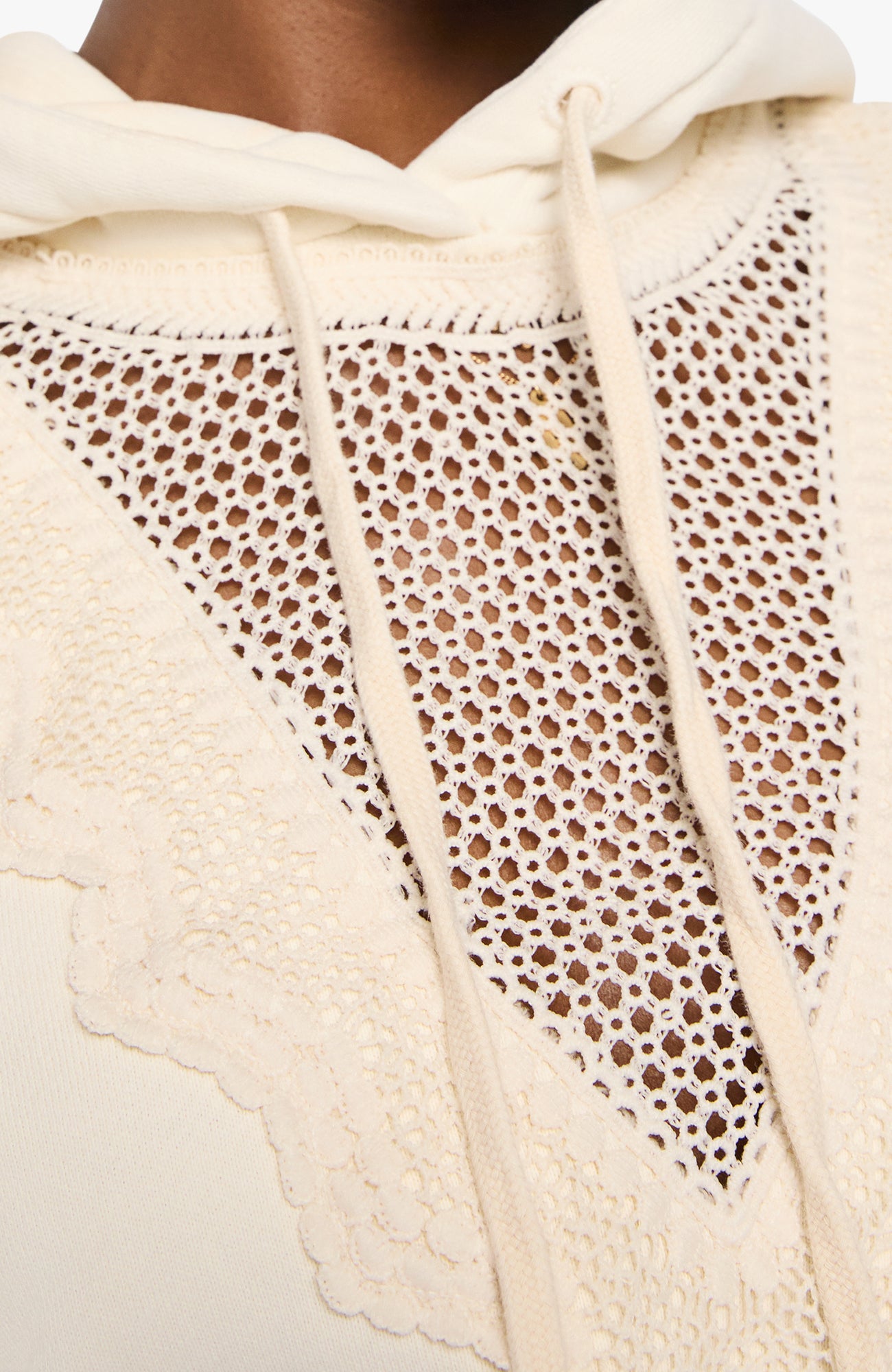 Close-up of a cream-colored knit hoodie featuring a large triangular open-knit lace panel on the front, with matching drawstrings and a scalloped edge.