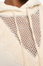 Close-up of a cream-colored knit hoodie featuring a large triangular open-knit lace panel on the front, with matching drawstrings and a scalloped edge.