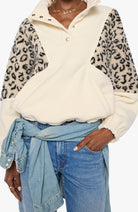 A person wearing a cream-colored quarter-zip pullover sweatshirt with leopard-print faux-fur sleeves, styled with blue jeans and a light blue denim shirt tied around the waist.