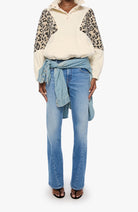 A model wearing a cream and leopard-print color-blocked pullover top, light-wash blue jeans with a light blue shirt tied around the waist, and black sandals, set against a plain white background.