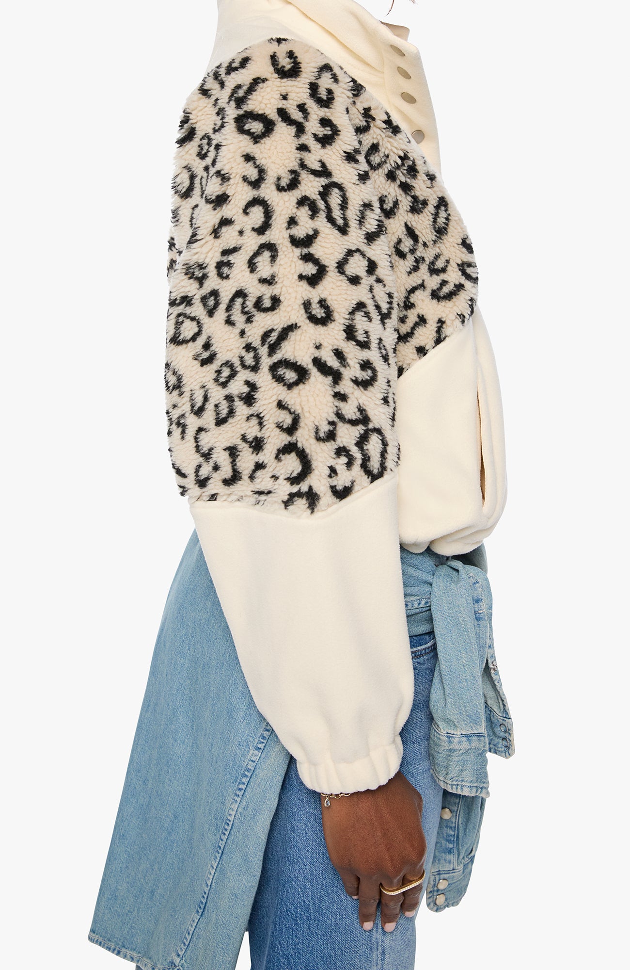 A cropped side view of a person wearing a fuzzy color-block cardigan with a beige and black leopard print on one half and solid cream on the other, paired with light blue denim jeans.