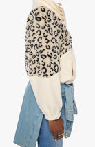 A cropped side view of a person wearing a fuzzy color-block cardigan with a beige and black leopard print on one half and solid cream on the other, paired with light blue denim jeans.