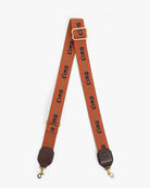 A wide orange-red fabric camera strap with a repeating black 'CIAO' logo print, featuring gold-tone metal hardware, a top adjustment buckle, and brown leather end caps with gold clasps, shown against a plain light gray background.