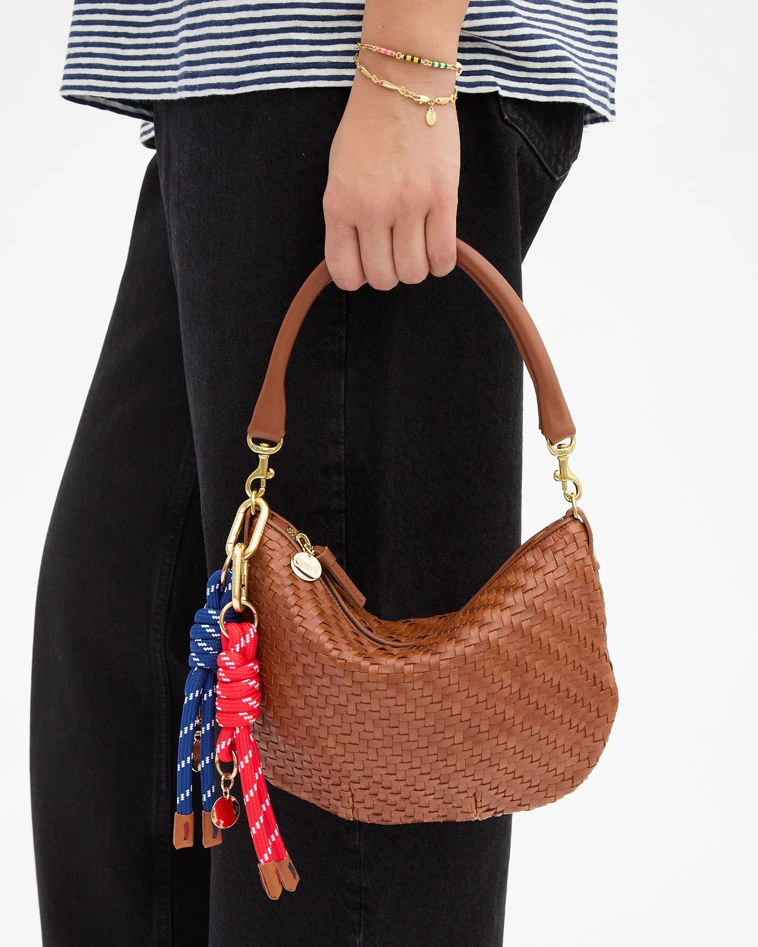 Brown woven handbag with colorful keychain held by a person wearing a striped shirt and black pants.