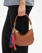 Brown woven handbag with colorful keychain held by a person wearing a striped shirt and black pants.