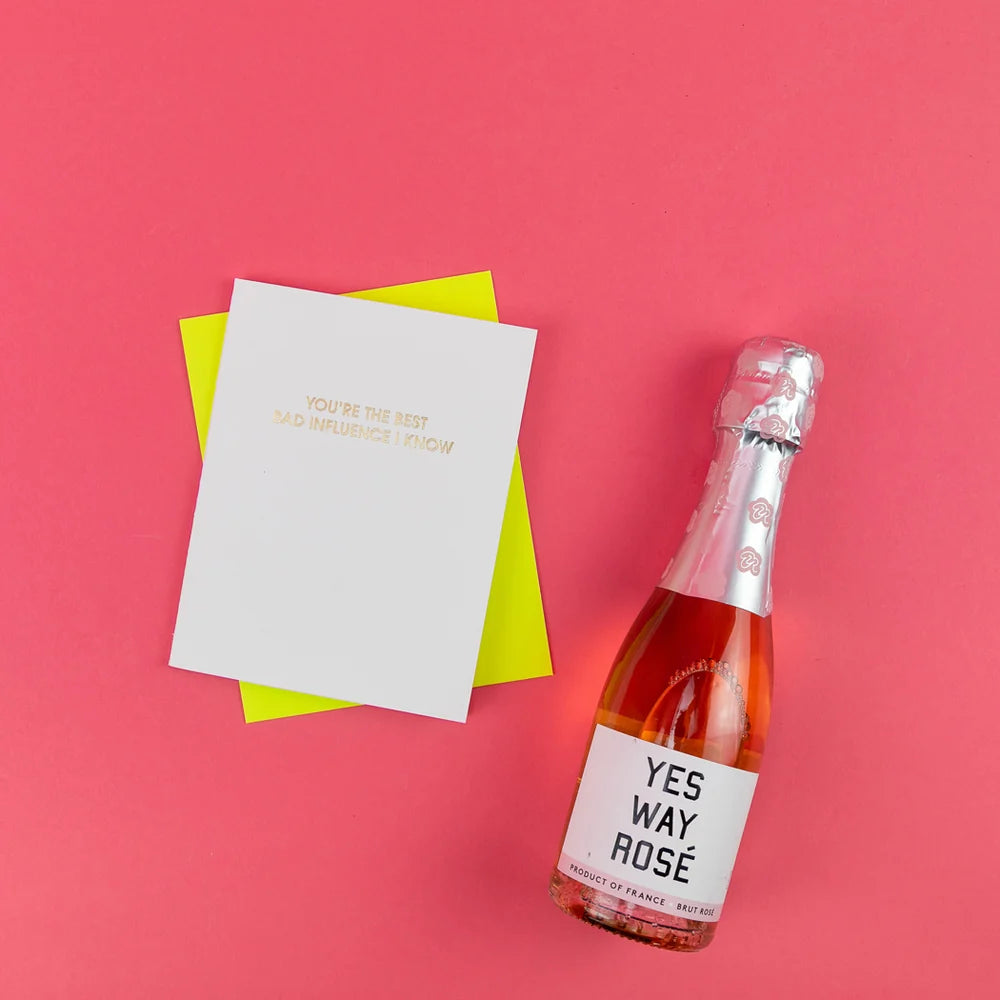 A white greeting card with gold text reading 'YOU'RE THE BEST SAD INFLUENCE I KNOW' lies on top of a bright yellow envelope, next to a bottle of rosé champagne with a white label that says 'YES WAY ROSÉ', all arranged on a solid pink background.