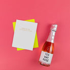 A white greeting card with gold text reading 'YOU'RE THE BEST SAD INFLUENCE I KNOW' lies on top of a bright yellow envelope, next to a bottle of rosé champagne with a white label that says 'YES WAY ROSÉ', all arranged on a solid pink background.