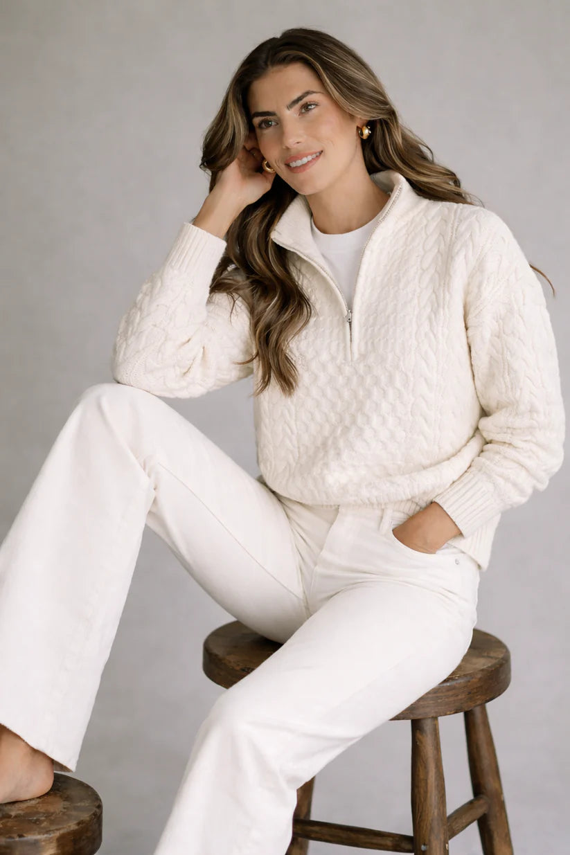 Woman wearing a white cable knit sweater and pants sitting on a wooden stool against a gray background
