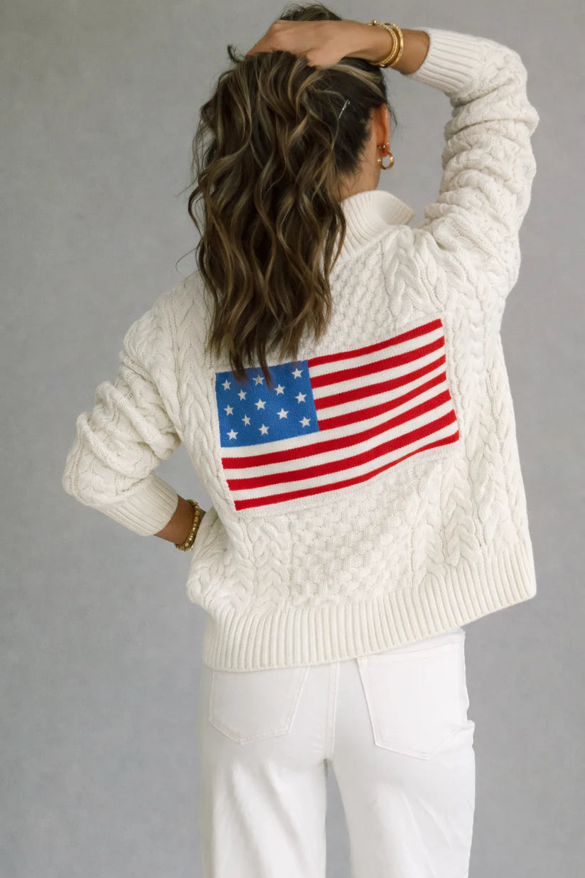 Person wearing a cream sweater with an American flag on the back against a gray background