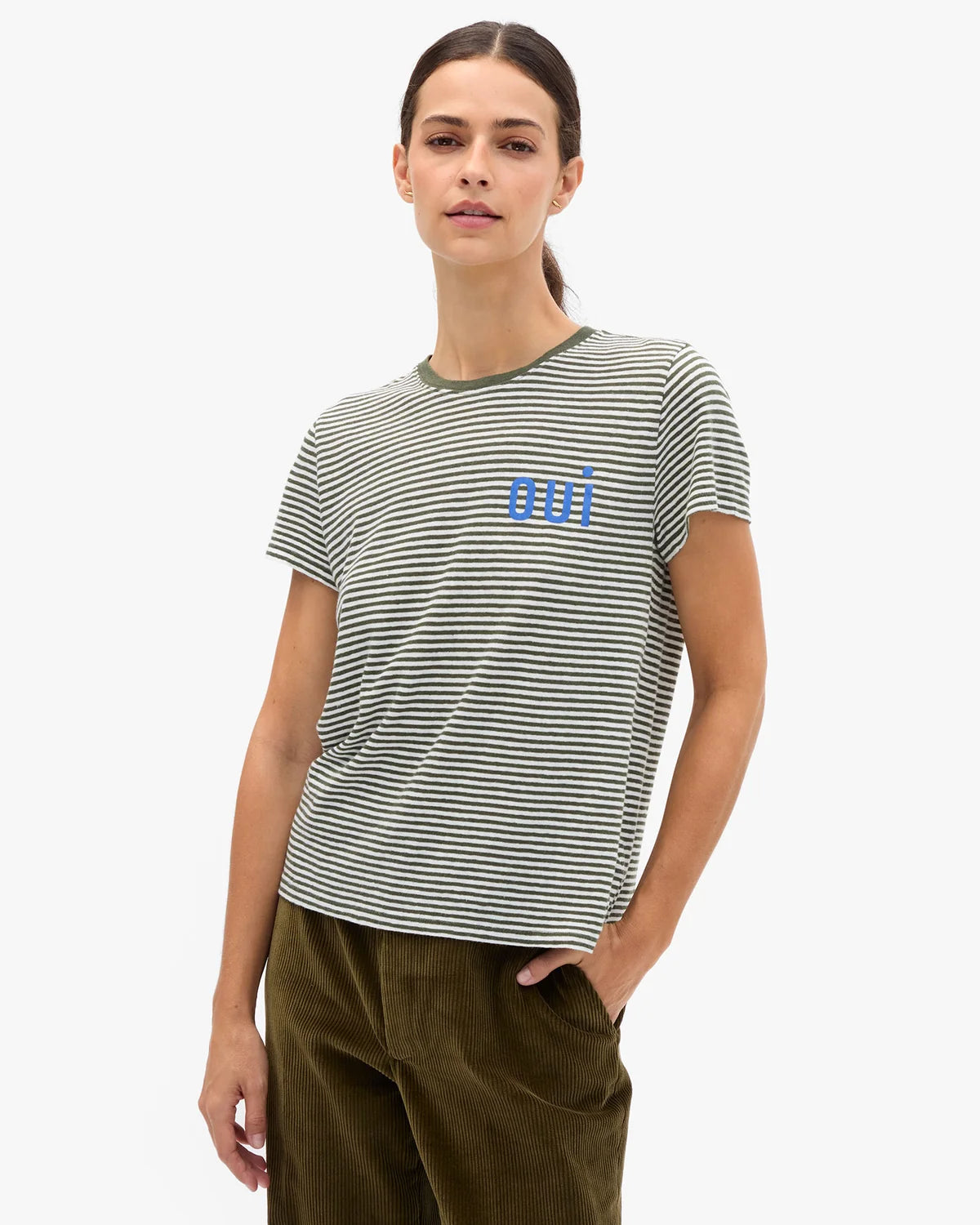 A woman modeling a short-sleeve crewneck t-shirt with thin horizontal black and olive green stripes and a blue 'mi' logo on the chest, paired with olive green pants against a light gray background.