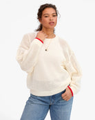 Woman wearing a cream sweater with red trim and blue jeans on a white background