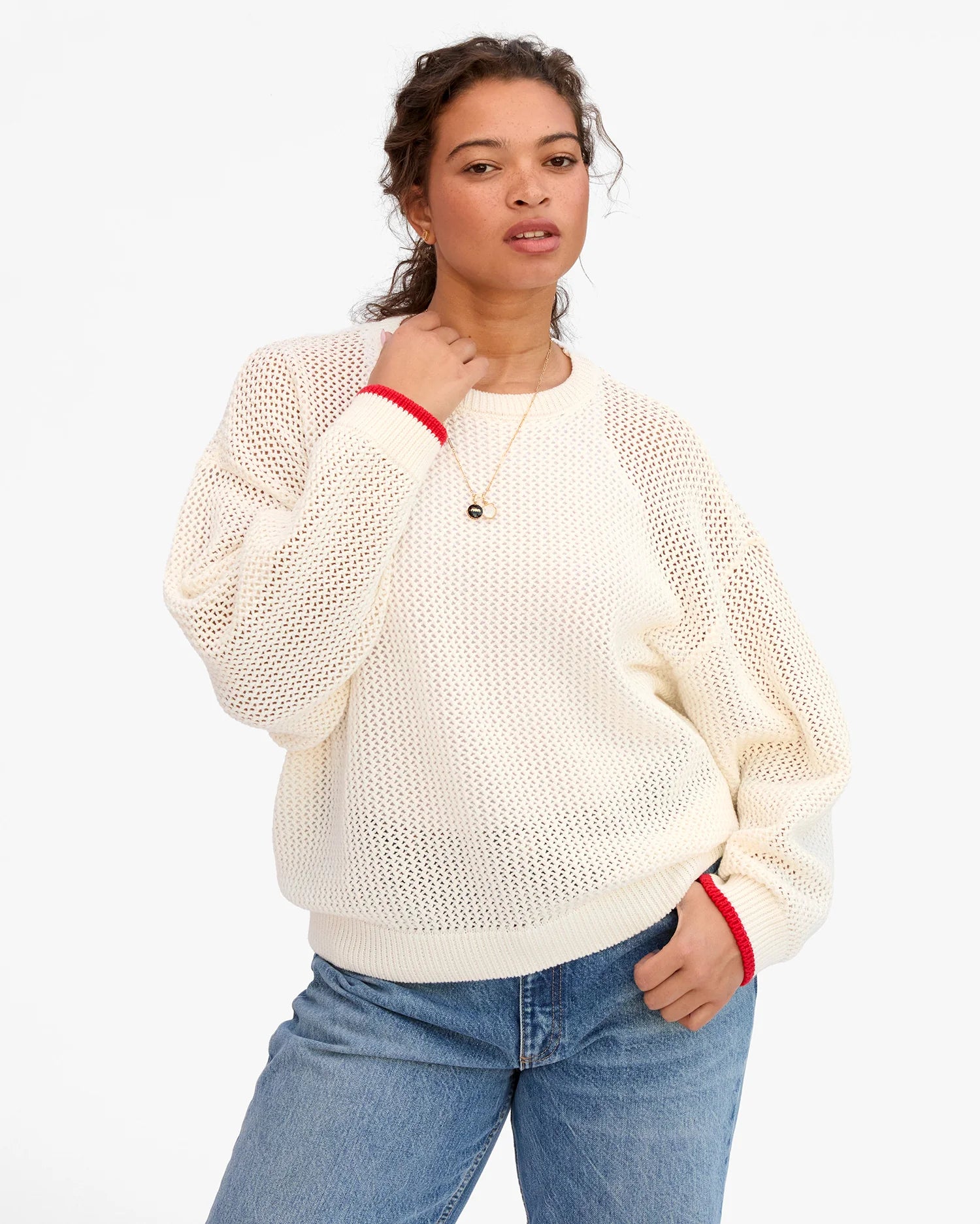 Woman wearing a cream sweater with red trim and blue jeans on a white background