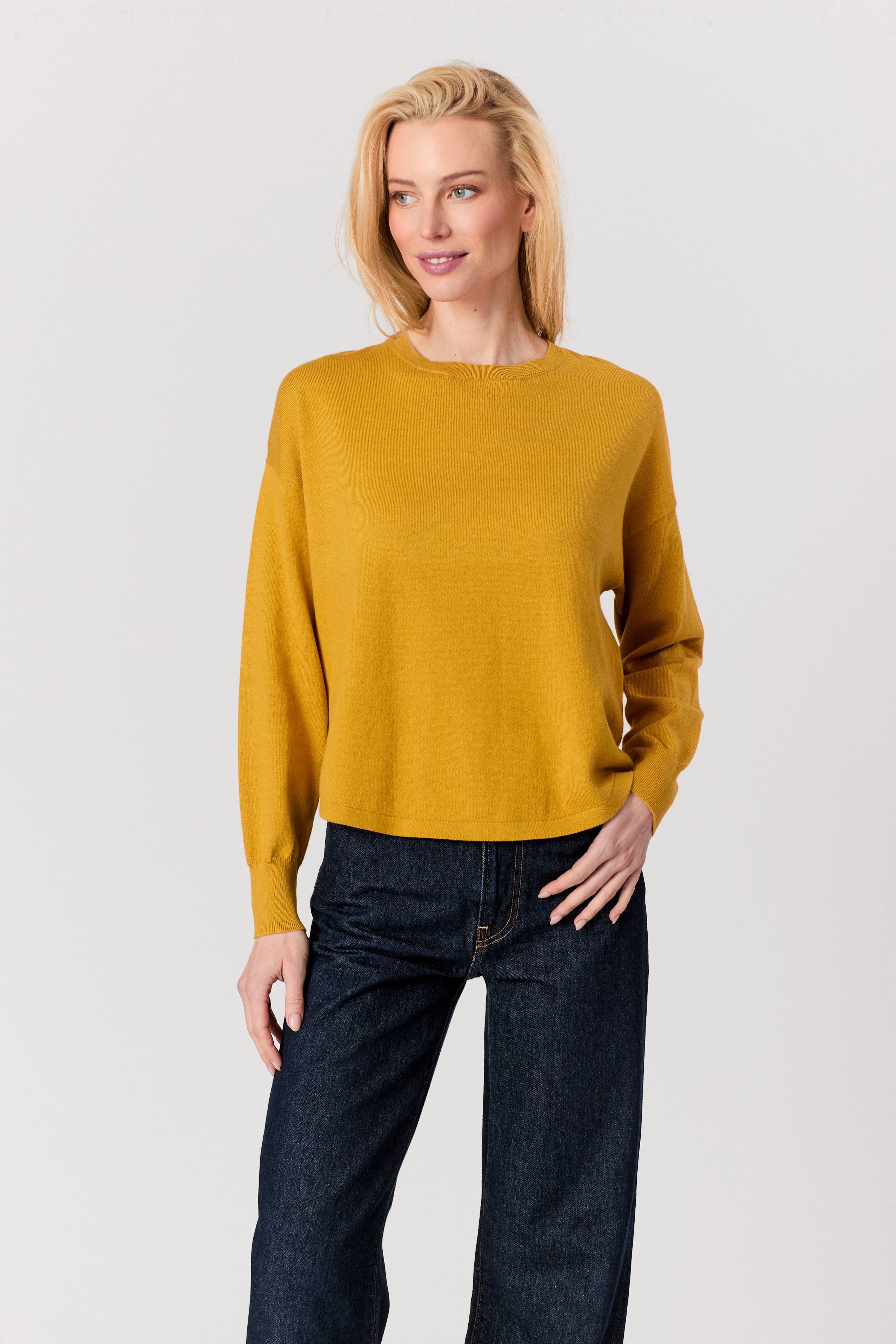 A blonde woman modeling a mustard yellow long-sleeve knit sweater with dark blue jeans against a light gray studio background.