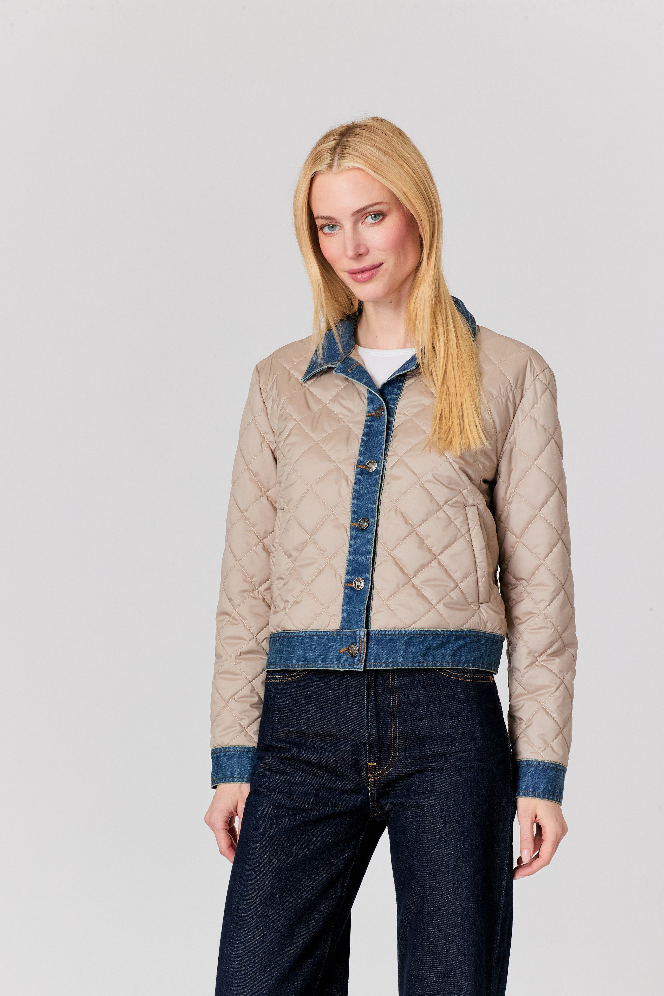 A blonde woman modeling a cropped quilted jacket with a beige body and blue denim trim, paired with dark blue jeans, against a solid light gray background.