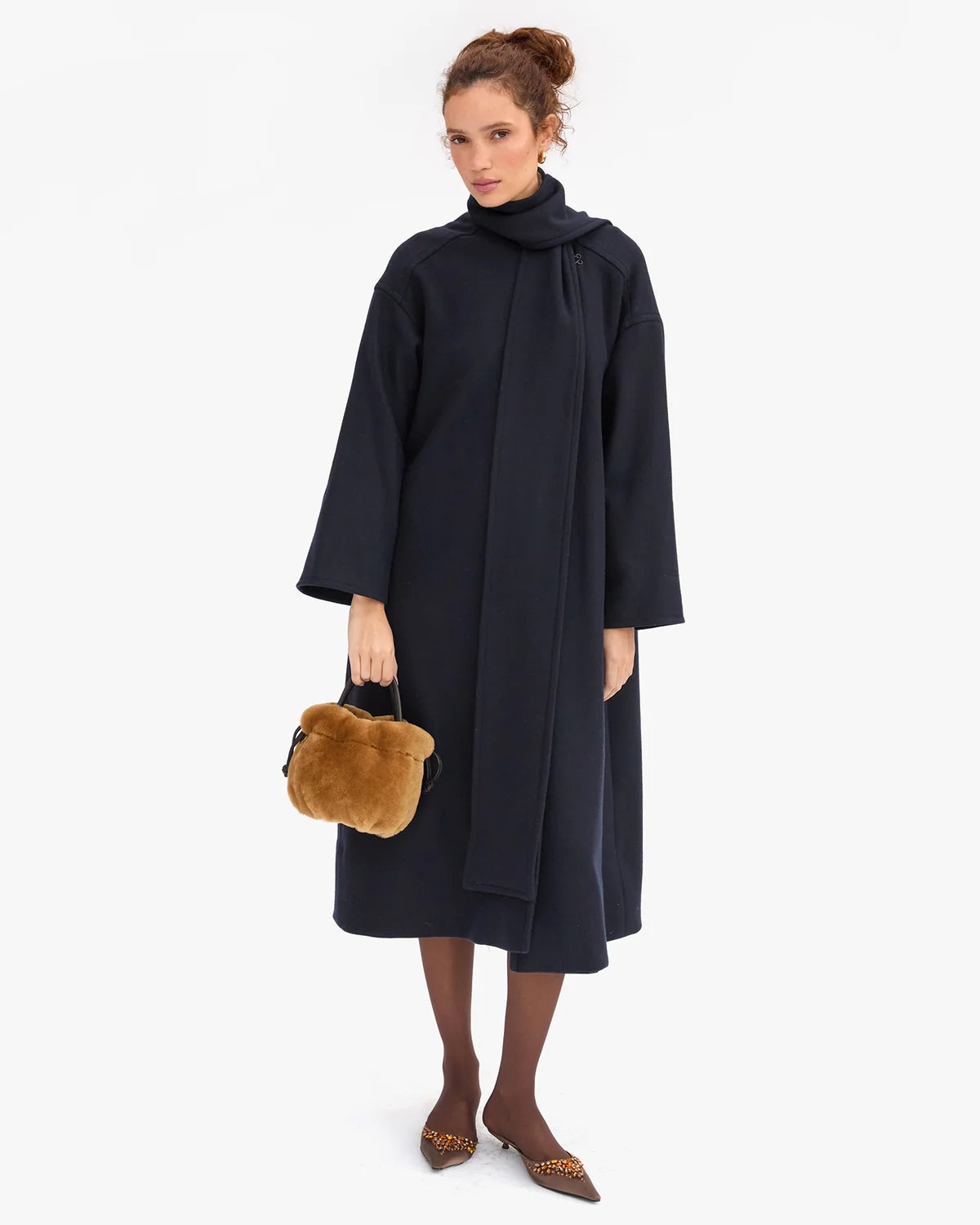 A model stands against a light gray studio background wearing a long, oversized dark navy wrap coat with wide sleeves and a high draped collar. She holds a small round brown fur handbag and wears brown pointed flats with leopard-print detailing.