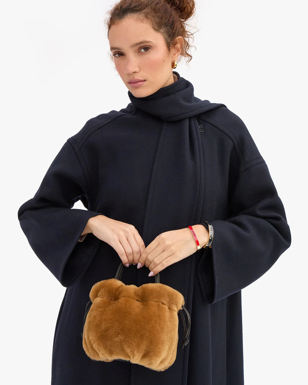 A woman wearing a dark navy blue coat with a high collar, holding a small brown plush handbag with a bear face design, against a plain white background.