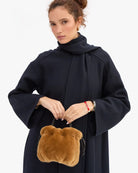 A woman wearing a dark navy blue coat with a high collar, holding a small brown plush handbag with a bear face design, against a plain white background.