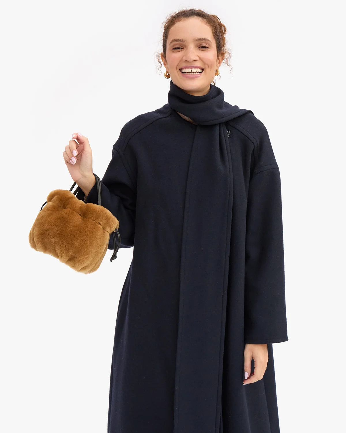 A woman wearing a long, dark navy wrap coat and holding a brown plush teddy bear handbag, photographed against a clean white studio background.