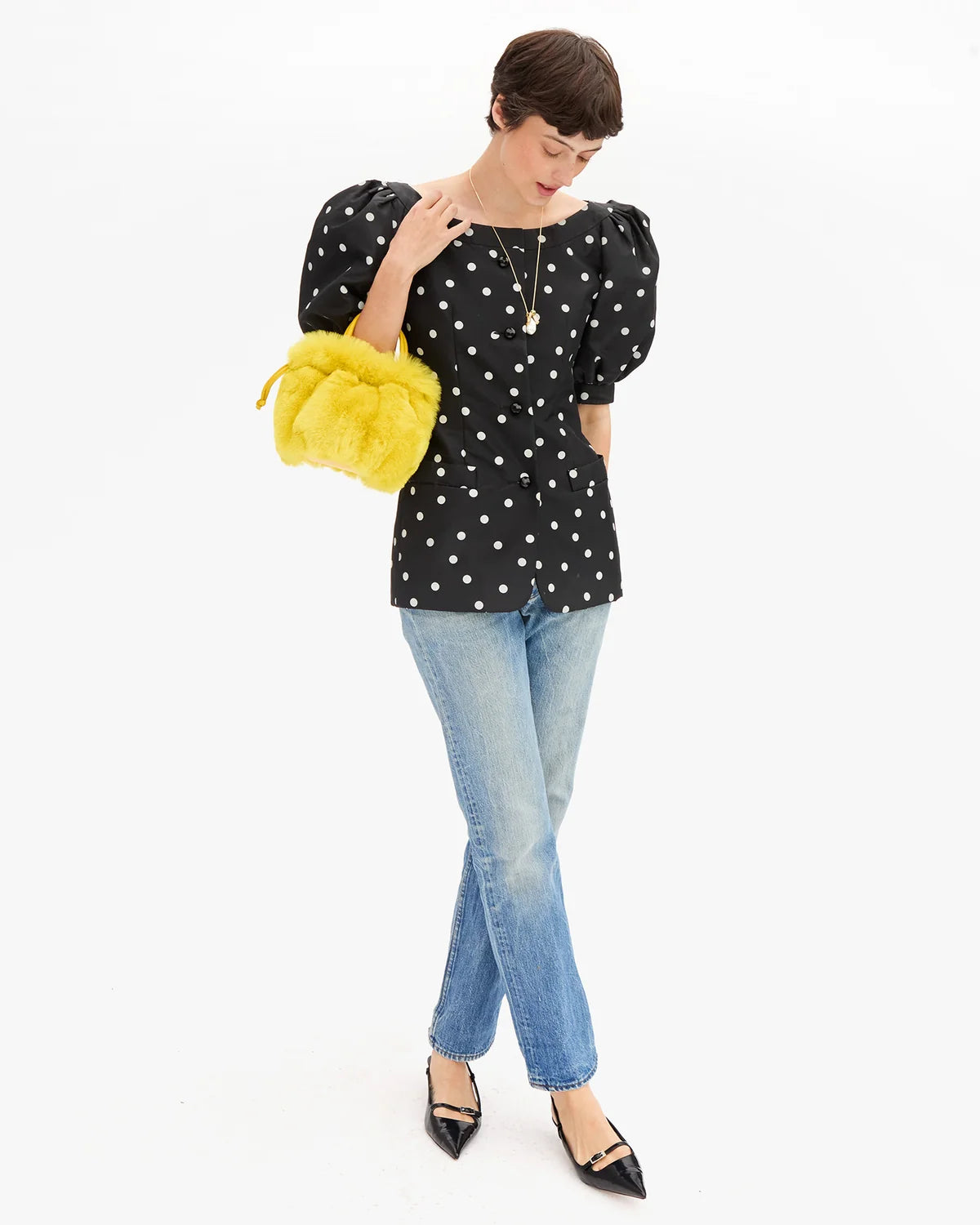 A woman modeling a black puff-sleeve blouse with white polka dots, paired with light-wash blue jeans, black pointed flats, and a bright yellow fuzzy crossbody bag, set against a plain white studio background.
