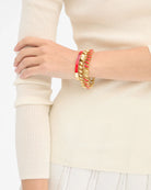 A close-up of a person's wrist wearing a stack of chunky gold chain and red striped bangles, paired with a cream ribbed knit sweater.