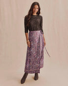A woman modeling a dark charcoal lace long-sleeve top and a purple patterned wrap midi skirt with brown ankle boots, standing against a warm beige studio backdrop.