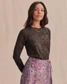A woman wearing a sheer black lace long-sleeve top and a purple floral patterned skirt, standing against a soft beige backdrop.