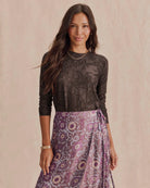 A woman modeling a dark brown lace long-sleeve top paired with a purple and pink paisley patterned wrap skirt, set against a warm beige textured background.