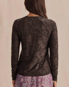 A woman viewed from the back wearing a dark brown sheer floral lace long-sleeve top and a purple patterned skirt, set against a plain beige background.