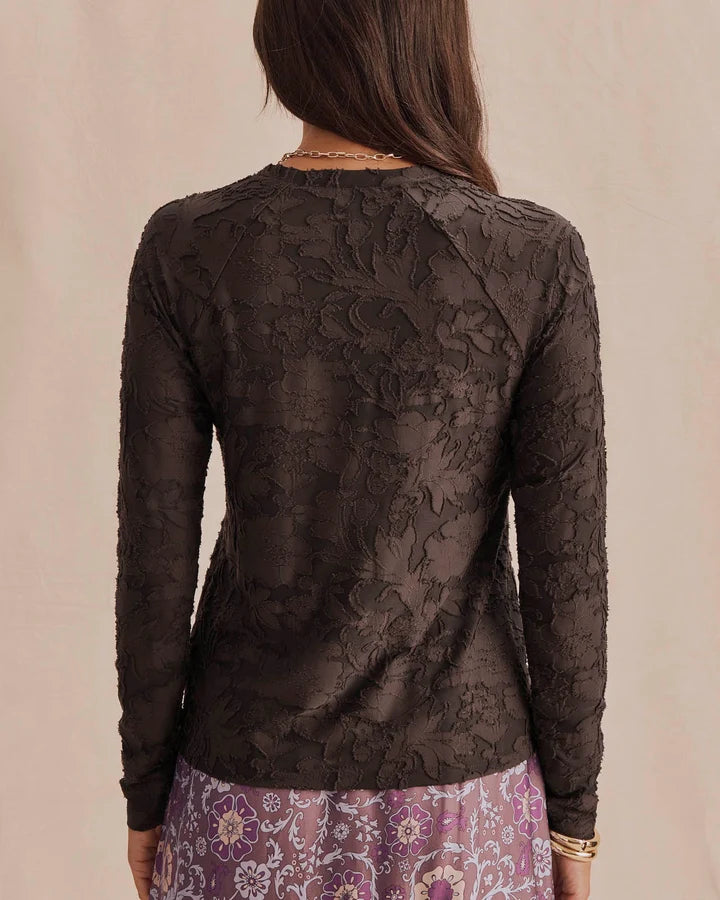 A woman viewed from the back wearing a dark brown sheer floral lace long-sleeve top and a purple patterned skirt, set against a plain beige background.