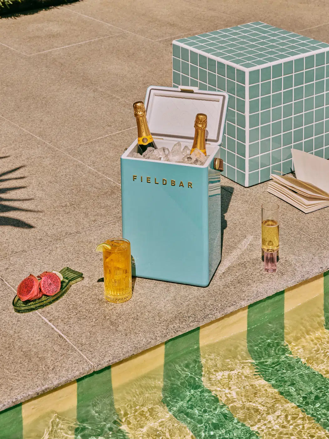 Blue cooler with 'FIELDBAR' branding next to a poolside setting with drinks and snacks.