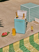 Blue cooler with 'FIELDBAR' branding next to a poolside setting with drinks and snacks.