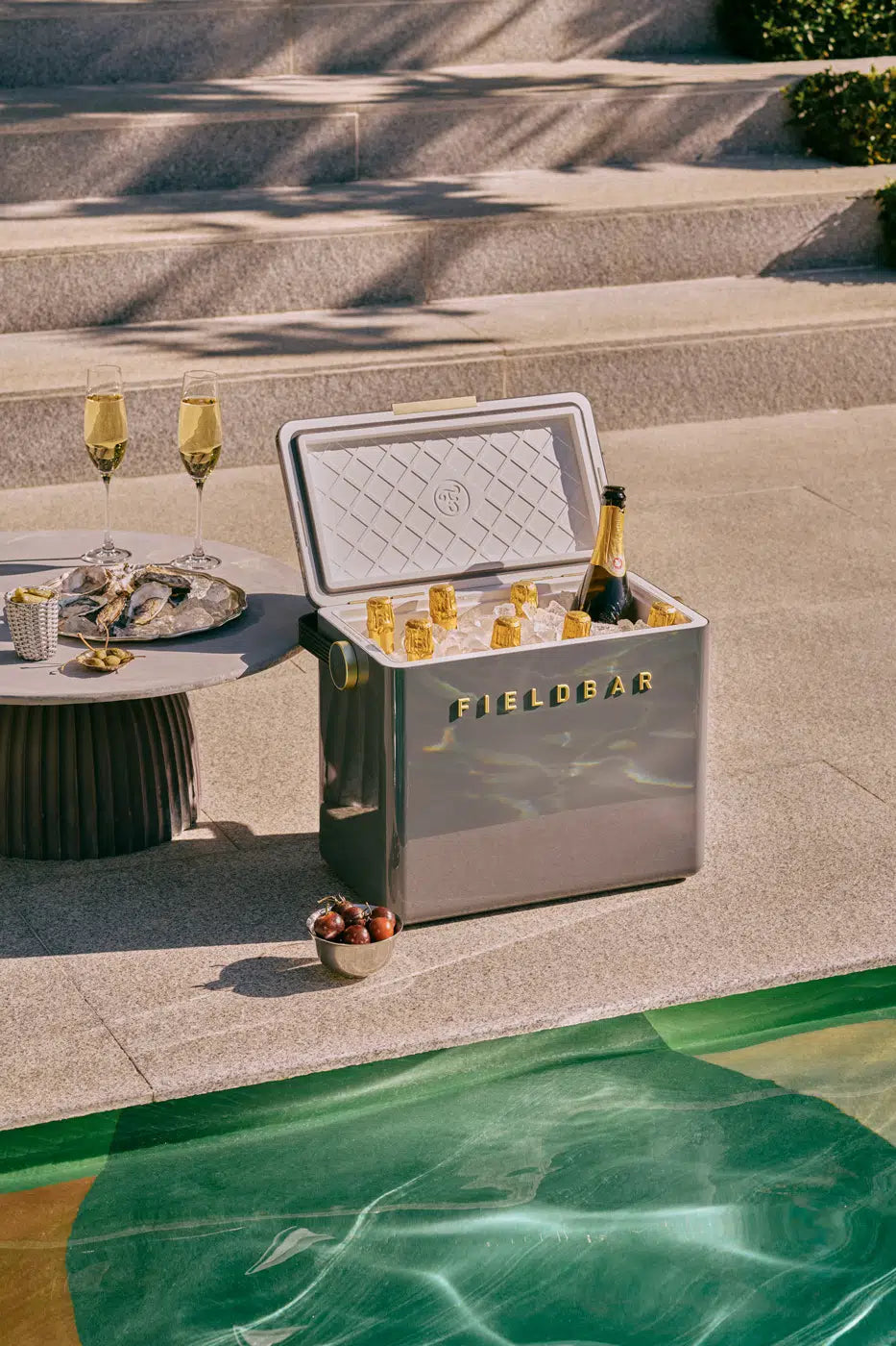 Gray cooler labeled 'FIELDBAR' with drinks by a poolside setting