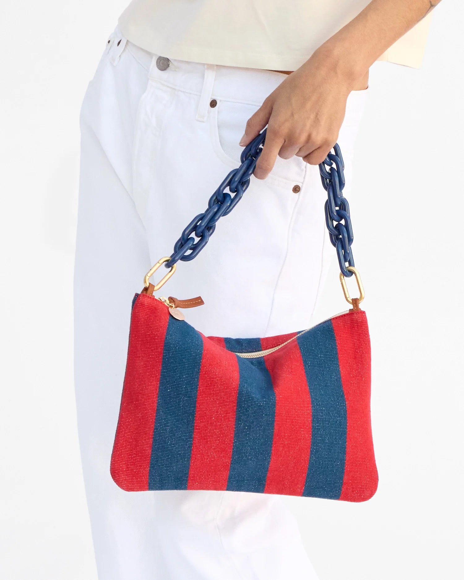 Red and blue striped handbag with a chain handle held by a person wearing white pants.