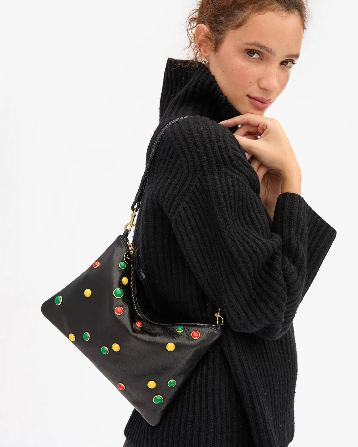 A woman wearing a black ribbed turtleneck sweater carries a black shoulder bag decorated with colorful green, yellow, and red studs, set against a plain white background.