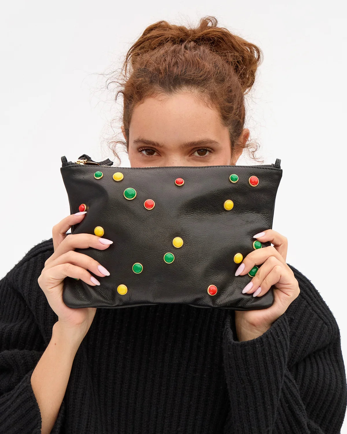 A woman holding a black faux-leather clutch bag decorated with colorful round studs in red, yellow, and green, posing with it against a plain white background.