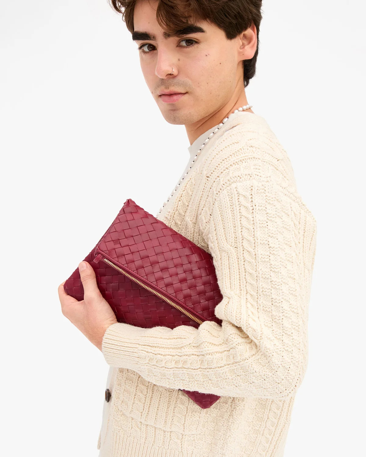 A man wearing a cream cable-knit sweater holds a burgundy woven leather clutch bag with a gold zipper against a plain white background.