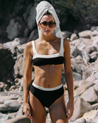 Woman in a black and white bikini with a towel on her head, standing on a rocky beach.