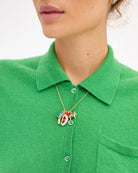A close-up of a model wearing a gold necklace with colorful enamel charms, styled with a bright green knit polo shirt against a clean white background.