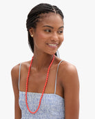 A smiling woman with braided hair wearing a long coral-red beaded necklace and a blue-and-white striped top, photographed against a plain light gray background.