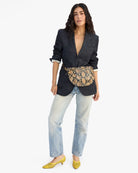 Woman wearing a dark blazer, light jeans, and yellow shoes with a snake print belt bag on a white background.
