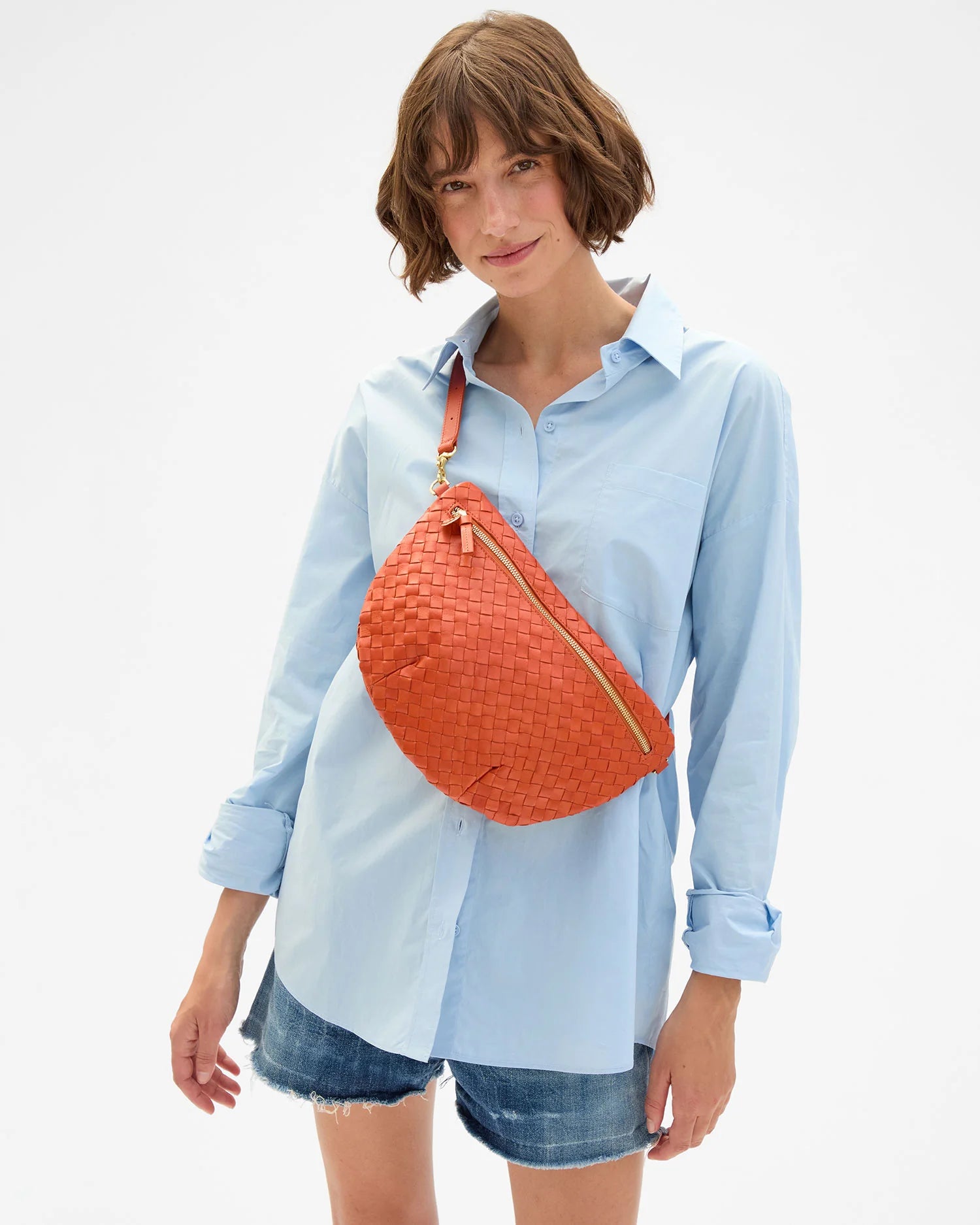 Woman wearing a light blue shirt and denim shorts with an orange crossbody bag on a white background