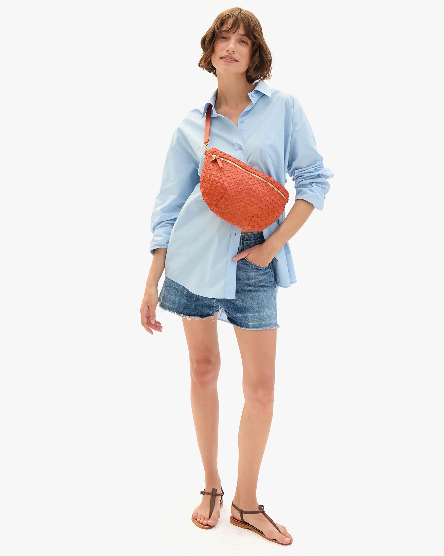 Woman wearing a light blue shirt, denim shorts, and carrying an orange bag on a white background