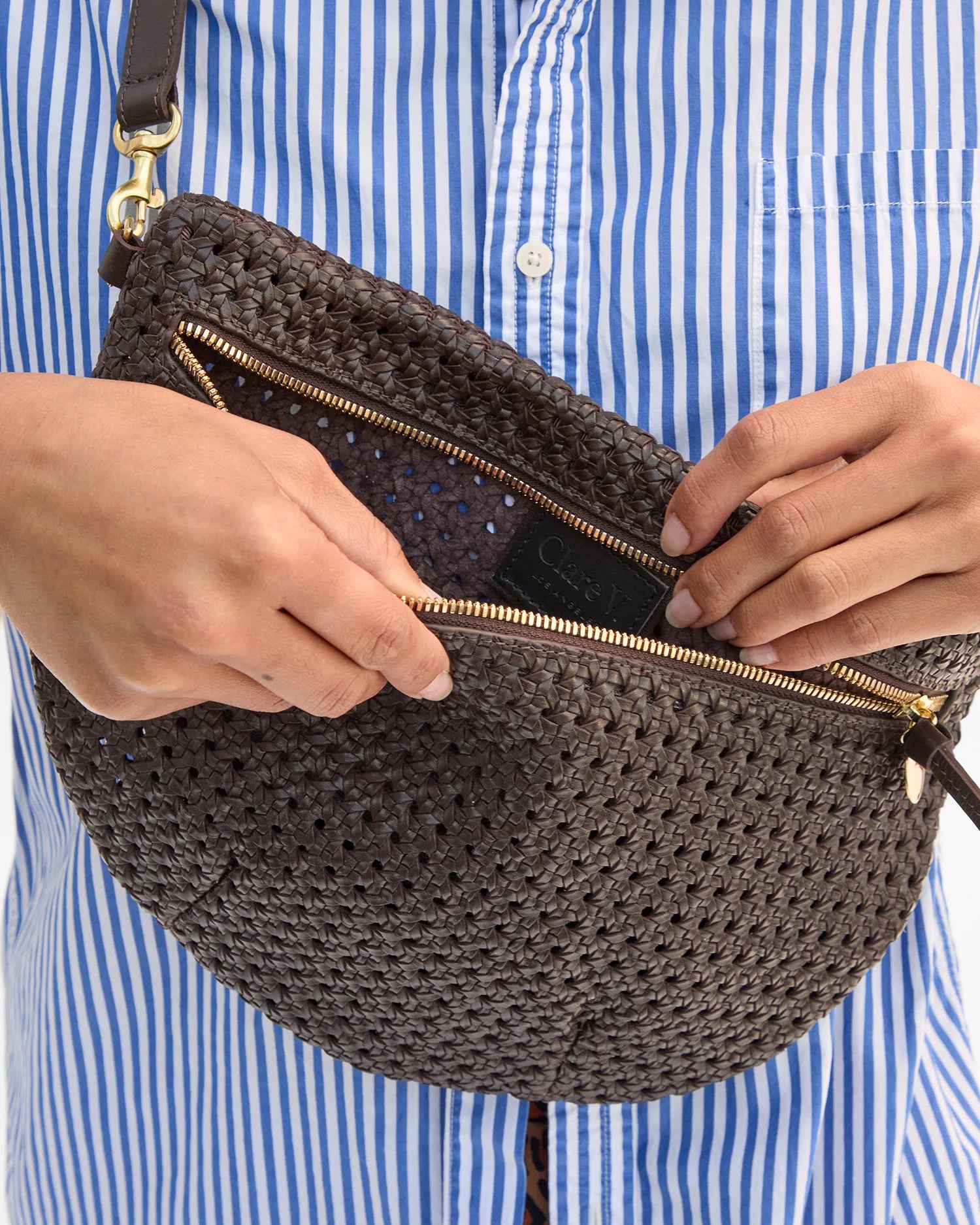 Person holding a brown woven handbag with a gold zipper against a blue and white striped shirt background.