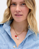 A close-up of a woman with blonde hair wearing a gold chain necklace with a white ceramic dog charm and red bead accents, paired with a light blue denim shirt.