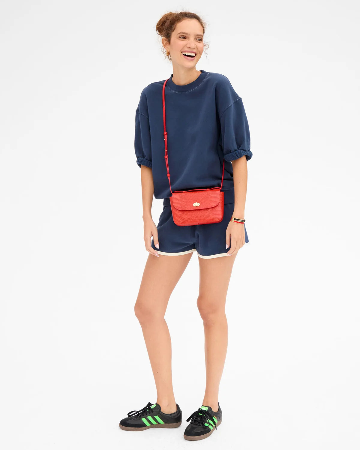 A woman modeling a navy blue sweatshirt and matching shorts, accessorized with a bright red textured crossbody bag with a gold clasp, against a plain white background.